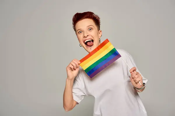 Modelo queer animado com piercing segurando pequeno LGBT plana e furando a língua no fundo cinza — Fotografia de Stock