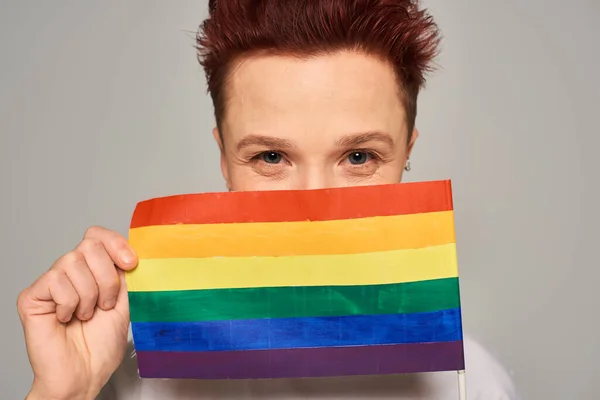 Fröhliche rothaarige queere Person, die ihr Gesicht mit einer kleinen LGBT-Fahne verdeckt und in die Kamera auf grau schaut — Stockfoto