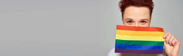 Joyful redhead queer person obscuring face with small LGBT flag looking at camera on grey, banner — Stock Photo