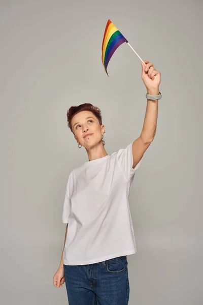 Redhead queer person in white t-shirt holding small LGBT flag in raised hand while standing on grey — Stock Photo