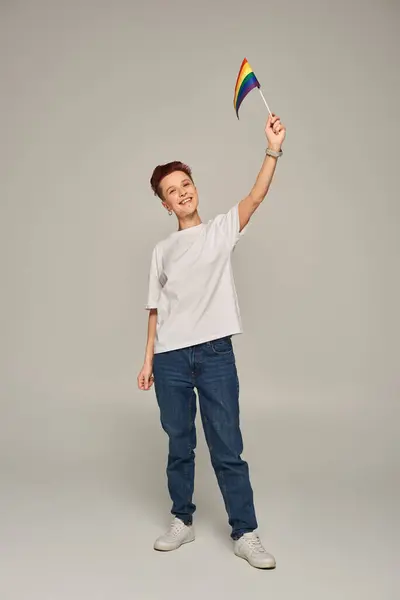 Cheerful queer person in white t-shirt holding small LGBT flag in raised hand while standing on grey — Stock Photo
