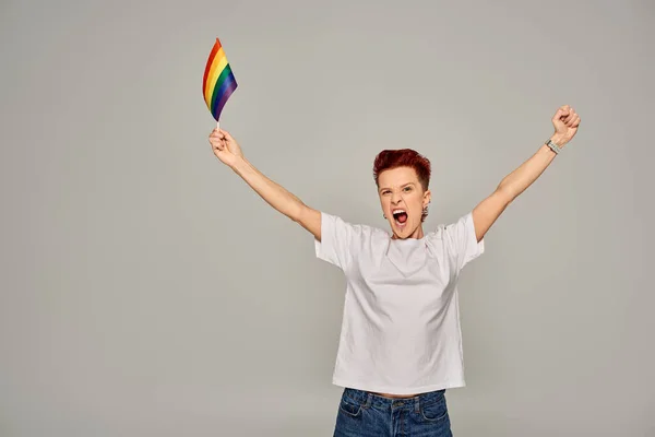 Emocionado persona queer en camiseta blanca de pie con pequeña bandera LGBT y gritando sobre fondo gris - foto de stock