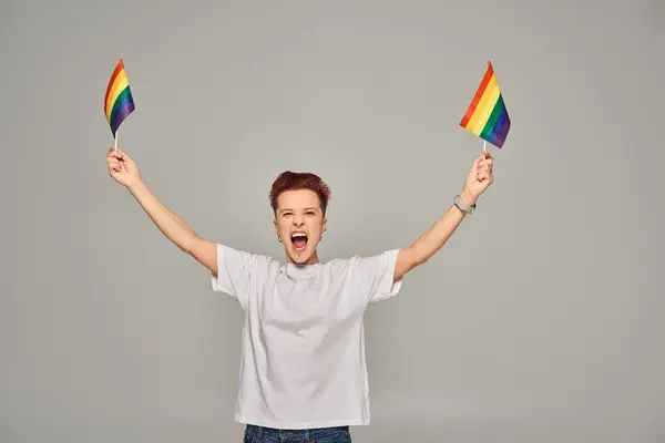 Persona queer eccitato in bianco t-shirt in piedi con piccola bandiera LGBT e urlando su sfondo grigio — Foto stock