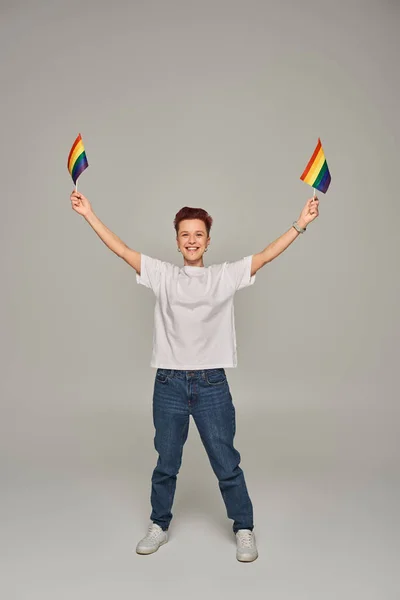 Fröhliche queere Person in weißem T-Shirt und Jeans, die mit kleinen LGBT-Fahnen in erhobenen Händen auf grau posiert — Stockfoto