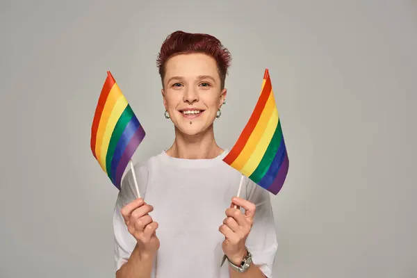 Joyful queer person in white t-shirt holding small LGBT flags and looking at camera on grey backdrop — Stock Photo