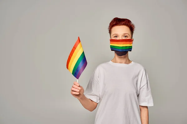 Pelirroja queer persona en blanco camiseta y arco iris colores máscara médica con pequeña bandera LGBT en gris - foto de stock