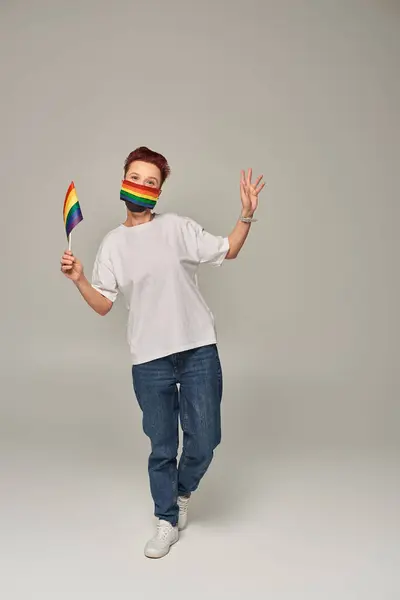 Intera lunghezza della persona queer rossa in colori arcobaleno maschera medica con piccola bandiera LGBT sul grigio — Foto stock