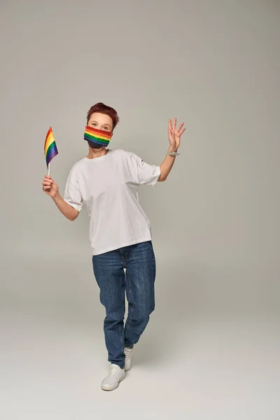 Full length of redhead queer person in rainbow colors medical mask with small LGBT flag on grey — Stock Photo