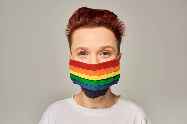 Porträt einer rothaarigen queeren Person in Regenbogenfarben mit medizinischer Maske, die vor grauem Hintergrund in die Kamera blickt — Stockfoto