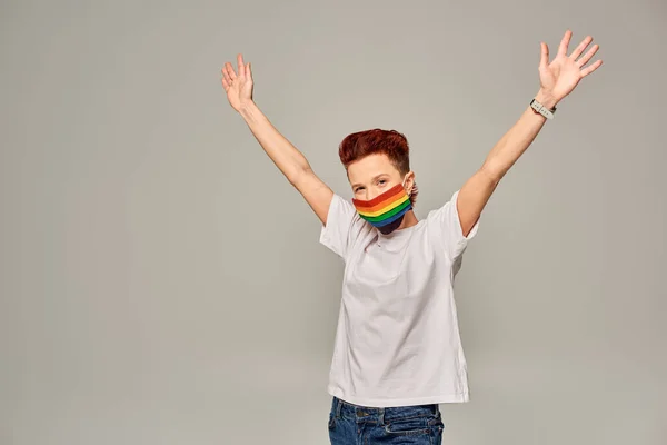 Redhead queer model in white t-shirt and rainbow colors medical mask with raised hands on grey — Stock Photo