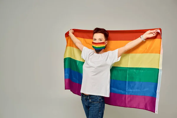 Modello queer rossa in t-shirt bianca e colori arcobaleno maschera medica con bandiera LGBT su grigio — Foto stock