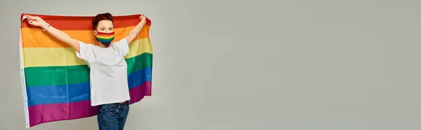 Руда квір-модель в веселкових кольорах медична маска, що тримає прапор ЛГБТ на сірому, горизонтальному банері — стокове фото