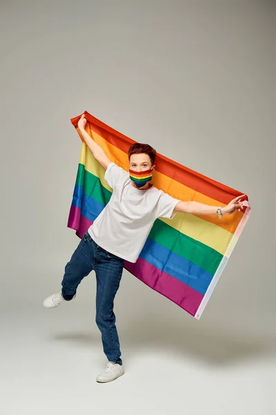Piena lunghezza del modello queer rossa in colori arcobaleno maschera medica che tiene bandiera LGBT sul grigio — Foto stock