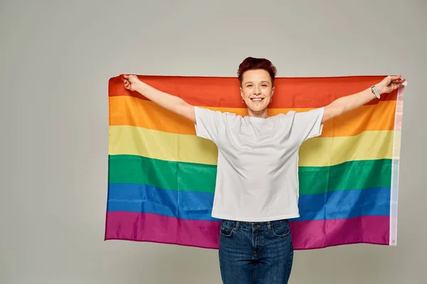 Cheerful redhead bigender person in white t-shirt standing with LGBT flag on grey backdrop — Stock Photo