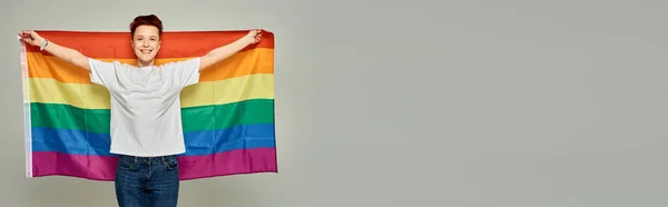 Alegre pelirroja persona grande en camiseta blanca de pie con bandera LGBT sobre fondo gris, bandera - foto de stock