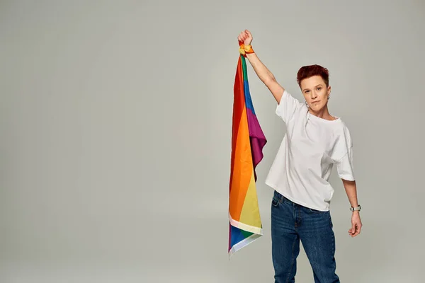 Serious redhead bigender person in white t-shirt and jeans standing with LGBT flag on grey backdrop — Stock Photo