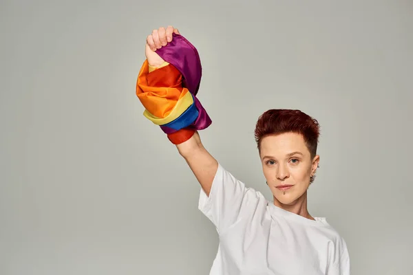 Confident redhead bigender person in white t-shirt and jeans standing with LGBT flag on grey — Stock Photo