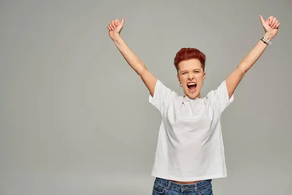 Excited non-binary person in white t-shirt showing thumbs up with raised hands and screaming on grey — Stock Photo