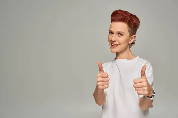 Happy redhead non-binary person in white t-shirt showing thumbs and smiling at camera on grey — Stock Photo