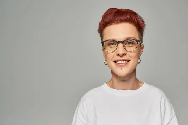 Portrait of happy redhead queer person in white t-shirt and eyeglasses looking at camera on grey — Stock Photo
