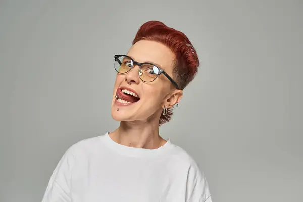 Funny and cheerful non-binary person in white t-shirt and eyeglasses sticking out tongue on grey — Stock Photo