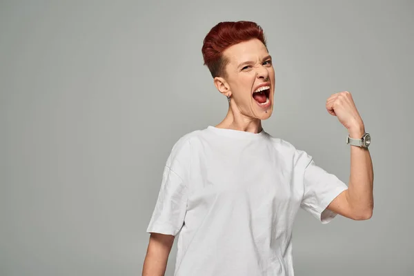 Esaltato rossa bigender persona in bianco t-shirt mostrando vincere gesto e urlando sul grigio — Foto stock