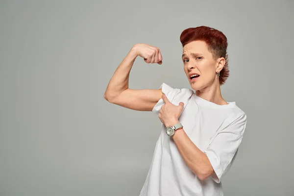 Displeased non-binary person in white t-shirt showing weak muscles and looking at camera on grey — Stock Photo
