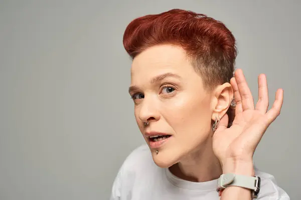 Curious redhead bigender person holding hand near ear while looking at camera on grey backdrop — Stock Photo