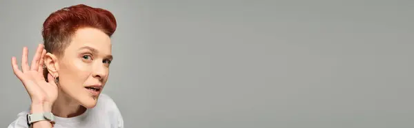 Curiosa rossa bigender persona che tiene la mano vicino all'orecchio mentre guarda la fotocamera sul grigio, banner — Foto stock