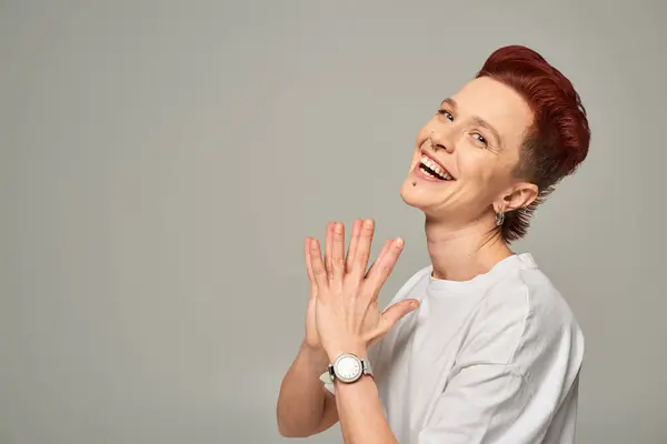 Cheerful redhead bigender person in white t-shirt laughing and looking at camera on grey, happiness — Stock Photo