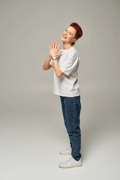 Cheerful redhead bigender person in white t-shirt and jeans laughing and looking at camera on grey — Stock Photo