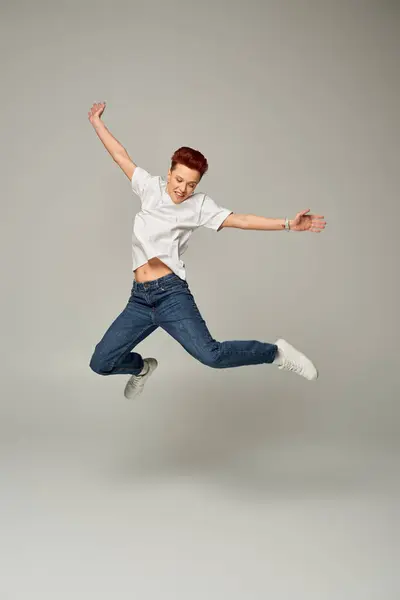 Full length of happy non-binary person in white t-shirt and jeans levitating on grey backdrop — Stock Photo