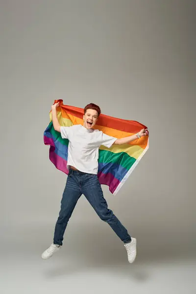 Persona queer muy alegre en camiseta blanca y pantalones vaqueros saltando y levitando con bandera LGBT en gris - foto de stock