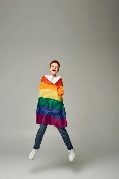 Persona queer muy alegre en camiseta blanca y pantalones vaqueros saltando y levitando con bandera LGBT en gris - foto de stock
