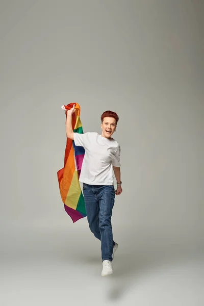 Überglückliche queere Person in weißem T-Shirt und Jeans und schwebend mit LGBT-Flagge auf grauem Hintergrund — Stockfoto
