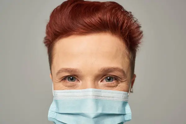 Primer plano retrato de pelirroja queer persona con feliz mirada en máscara médica mirando a la cámara en gris - foto de stock