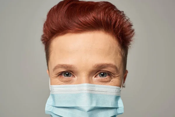 Close up portrait of redhead queer person with happy gaze in medical mask looking at camera on grey — Stock Photo