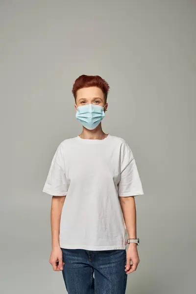 Positivo rossa queer persona vincere bianco t-shirt e maschera medica in piedi su sfondo grigio — Foto stock