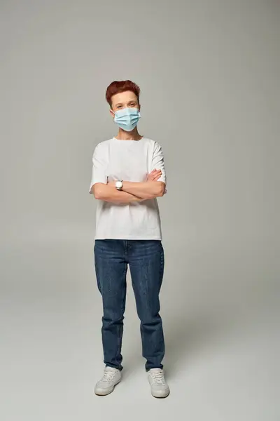 Positive rothaarige queere Person gewinnt weißes T-Shirt und medizinische Maske stehend mit verschränkten Armen auf grau — Stockfoto