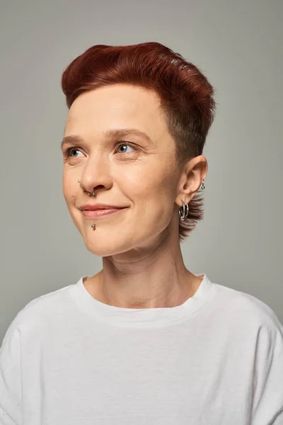 Portrait of redhead non-binary model with facial piercing smiling and looking away on grey — Stock Photo