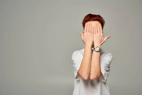Redhead bigender person in white t-shirt obscuring face with hands while standing on grey backdrop — Stock Photo