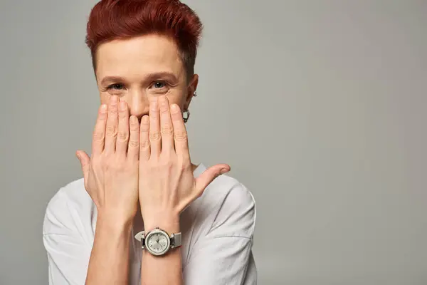 Redhead bigender person with cheerful gaze obscuring mouth with hands and looking at camera on grey — Stock Photo