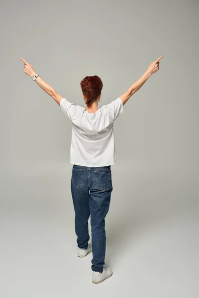 Back view of redhead queer person in white t-shirt and jeans pointing aside with fingers on grey — Stock Photo