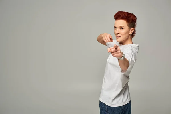 Pelirroja positiva queer persona en camiseta blanca y jeans apuntando con los dedos a la cámara en gris - foto de stock