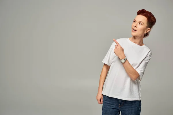 Thoughtful redhead non-binary person in white t-shirt looking away and pointing with finger on grey — Stock Photo