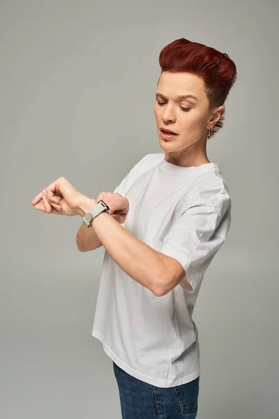 Serious redhead bigender person in white t-shirt checking time on wristwatch on grey backdrop — Stock Photo