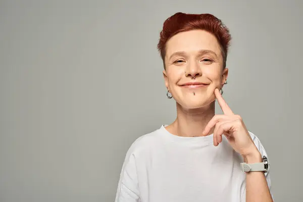 Joyful redhead bigender person in white t-shirt pointing at cheek and asking for kiss on grey — Stock Photo