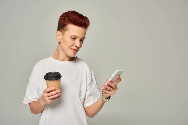 Sonriente pelirroja queer persona sosteniendo café para ir en taza de papel y mirando en el teléfono inteligente en gris - foto de stock