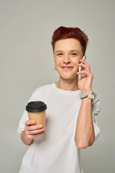 Smiling redhead queer person holding coffee to go in paper cup and talking on smartphone on grey — Stock Photo