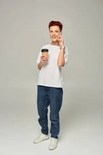 Smiling redhead queer person holding coffee to go in paper cup and talking on smartphone on grey — Stock Photo